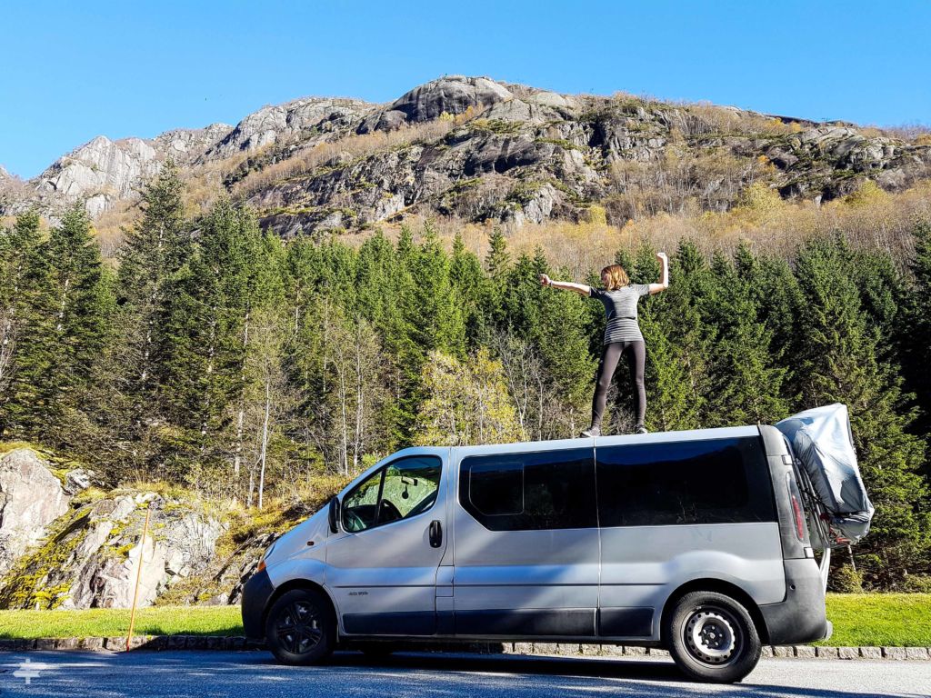 Mujer subida encima de una furgoneta con árboles y una montaña de fondo, en Noruega